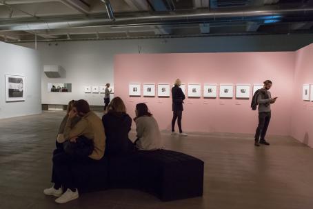 Woodmanin valokuvia esillä näyttelyssä, näyttelykävijöistä osa seisoo katsomassa kuvia ja osa istuu keskellä olevalla penkillä.