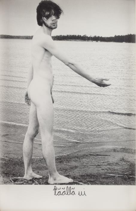 Mustavalkoisessa kuvassa alaston mies seisoo järven rannalla selin, hän katsoo kameraan päin ja viittaa toisella kädellään veteen. Alareunassa on teksti: "täällä ui".