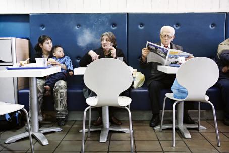 Kolme pientä valkoista pöytää vierekkäin. Vasemmalla pöydässä istuu nuori nainen vauva sylissään, keskellä vanhempi nainen ja oikeassa reunassa vanhempi mies lukee lehteä.