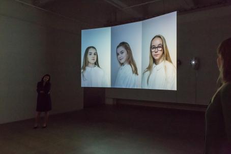 Näyttelytilassa oleva teos jossa on vierekkäin kolme muotokuvaa, joissa olevilla nuorilla on valkoiset albat päällään.