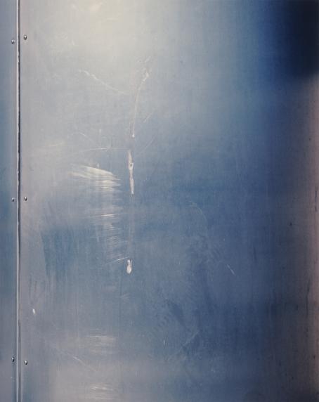 Andrei Lajunen: Seinä, noin 1997-1998, mustesuihkutuloste, 47 x 37 cm. Andrei Lajusen rahaston editio 10. Suomen valokuvataiteen museo. Siniharmaasävyinen, metalliselta näyttävä sileä pinta, jossa on vähän naarmuja. Vasemmalla on pystysuora sauma, jonka molemmin puolin on pieniä pyöreitä ruuvinkohtia.