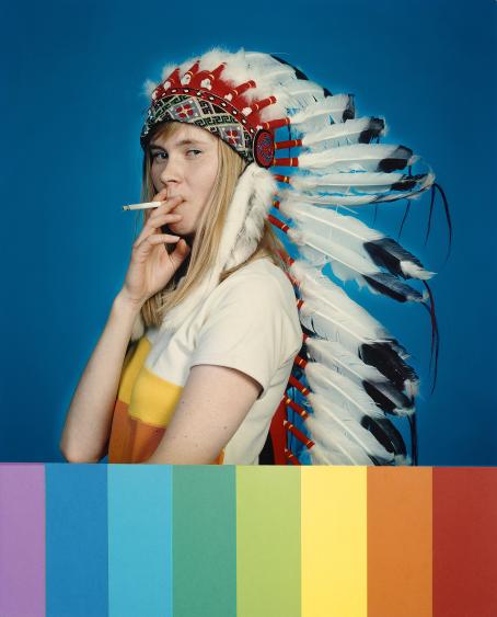 Nuorella naisella on päässään sulkapäähine. Hän polttaa tupakkaa ja katsoo sivusilmällä kameraan. Kuvan alareunassa on sateenkaaren värit pystyviivoina. 