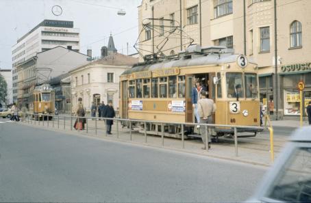 Haaleanruskea raitiovaunu numero 3 on pysähtynyt pysäkille. Vähän matkaa sen takana tulee raitiovaunu numero 2. Takana näkyy rakennuksia.