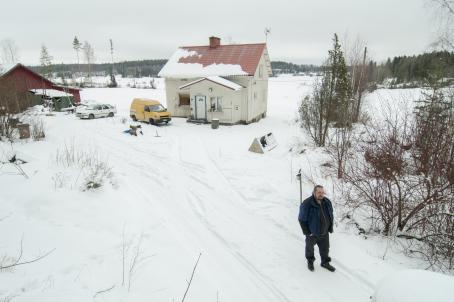 Ilmasta otettu kuva, jossa mies seisoo lumisella pihatiellä. Miehen takana on pieni vaalea omakotitalo, jonka pihassa on kaksi autoa ja kauempana punainen varastorakennus. Talon takana on peltoa ja kauempana metsää. 