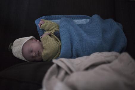 Pieni vauva vihreässä neulepaidassa ja valkoisessa neulepipossa nukkuu. Vauvan päällä on sininen neuleviltti. 