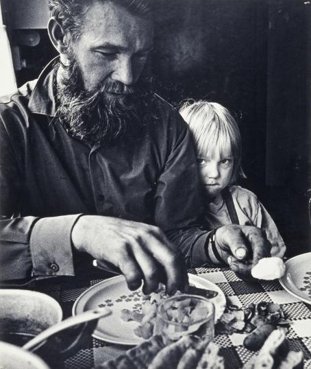 Mustavalkoisessa kuvassa parrakas mies istuu ruokapöydässä ja kuorii perunaa. Miehen vieressä istuu lapsi ja katsoo ujosti suoraan kameraan miehen takaa. 