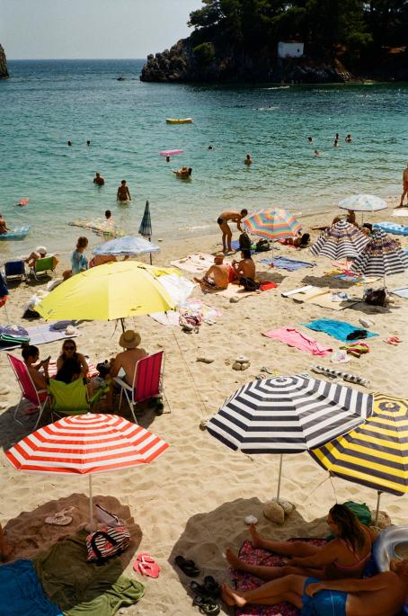 Aurinkoinen päivä hiekkarannalla. Rannalla on paljon ihmisiä istumassa vilttien päällä tai tuoleilla, ja monella on aurinkovarjo pystyssä. Vedessä on paljon uimareita.