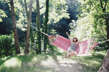 Epätarkka kuva, jossa nuori nainen istuu kahden puun väliin viritetyssä riippumatossa. Naisella on kädet ylhäällä, aurinkolasit päässä ja suu auki. Aurinko paistaa, ympärillä on vihreää metsää. 