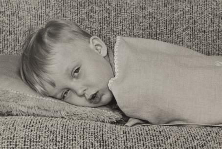 Mustavalkoisessa kuvassa pieni poika makaa sohvalla viltin alla. Pojan silmät ovat melkein puoliksi kiinni ja suu hieman auki. 