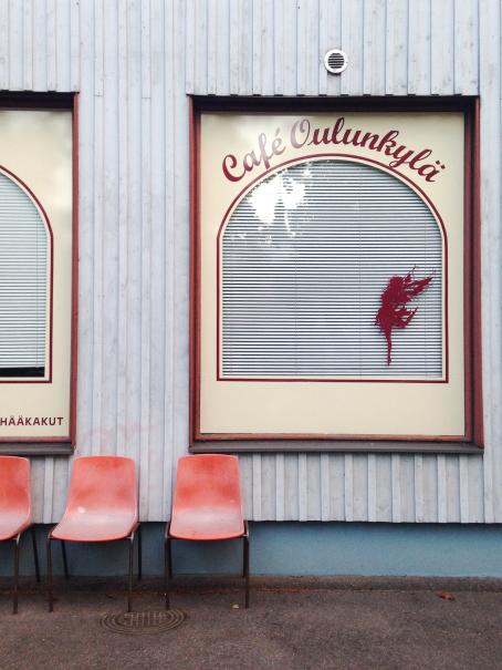 Rakennuksen seinä, jonka edessä on punaisia muovituoleja rivissä. Rakennnuksen ikkunassa lukee Cafe Oulunkylä.