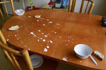 Punaruskealla pöydällä on kaksi tyhjää kulhoa, leivänpalasia ja jogurtinroiskeita. Tuolilla on tyhjä lautanen.