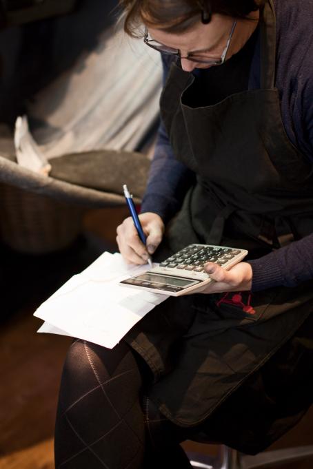Nainen istuu laskin kädessä ja kirjoittaa jotain jalkansa päällä olevaan paperiin. Naisella on musta essu päällä.