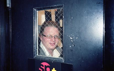 Tummassa ovessa on verkkopäällysteinen ikkuna, jonka takana näkyy miehen kasvot. Miehellä on taakse vedetyt hiukset, silmälasit ja kauluspaita.