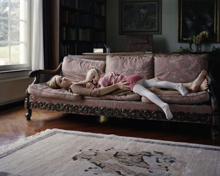 Koristeellisella sohvalla makaa selällään nainen. Osittain naisen päällä makaa mahallaan pieni tyttö, jolla on vaaleanpunainen mekko ja letit. Sohvan päädyssä näkyy ikkuna, sohvan takana hämärässä näkyy kirjahylly ja tauluja seinällä. 