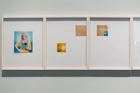 Seinällä on vierekkäin kolme vaaleaa kehystä, joiden sisällä on valkoisella taustalla pieniä kuvia. Vasemman reunan kuvassa nainen pitää kädessään paperia, jossa on erivärisiä neliöitä. Keskellä on kaksi pientä neliönmuotoista kuvaa, toinen on keltasävyinen ja toinen oranssiin vivahtava. Oikeanpuolimmaisessa on yksi pieni neliön muotoinen kuva, jossa näkyy seinää ja vähän ikkunaa.