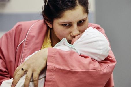 Nuori nainen vaaleanpunaisessa sairaalapaidassa pitää sylissään vauvaa ja antaa hänelle suukon kasvoille.