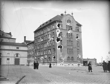 Mustavalkoisessa kuvassa korkea talo, jossa on seinässä isoja reikiä. Talon edessä on aukio, jossa kävelee ihmisiä. 