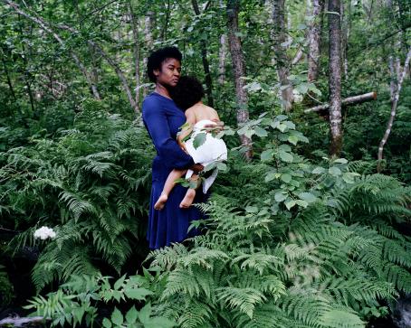 Tummaihoinen nainen seisoo vihreässä metsässä saniaisten keskellä ja pitää sylissään lasta, jonka vyötärön ympärillä on valkoinen kangas. Naisella on sininen mekko, ja sekä hänellä että lapsella on mustat afrohiukset.