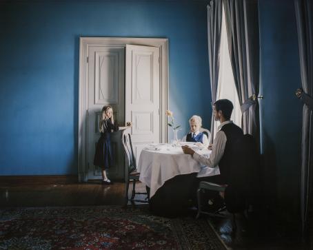 Siniseinäisessä huoneessa mies ja poika istuvat valkoisella pöytäliinalla peitetyn pyöreän pöydän ääressä. Heillä on kauluspaidat ja solmiot. Takimmaisella seinällä on ovi, jota nuori tyttö sinisessä mekossa on avaamassa. Hän katsoo kameraan, mies katsoo tyttöä. 