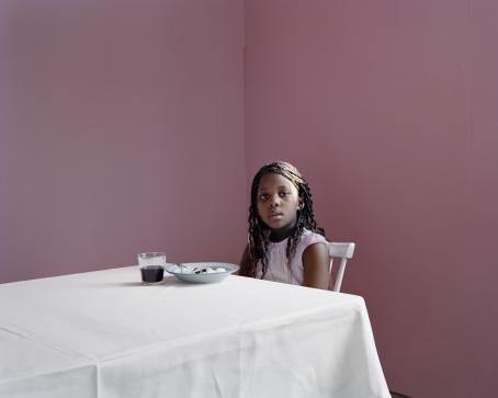Tummaihoinen tyttö istuu valkoisella pöytäliinalla peitetyn pöydän ääressä ja katsoo kameraan. Pöydällä on ainoastaan lautanen, jossa on jotain ruokaa, sekä lasi puolillaan jotain tummahkoa nestettä. Takana oleva seinä on roosan sävyinen.