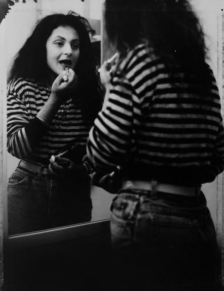 Mustavalkoisessa kuvassa raitapaitainen, tummatukkainen nainen seisoo peilin edessä levittämässä huulipunaa. Naisen kasvot näkyvät peilin kautta. 
