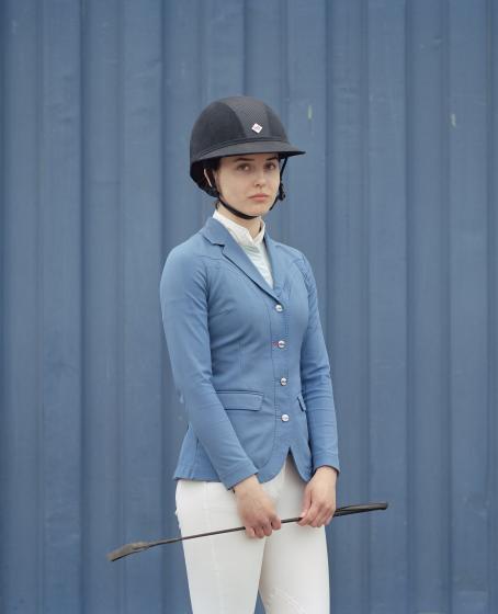 Nuori nainen sinisessä takissa ja valkoisissa housuissa seisoo sinisen lautaseinän edessä. Hänellä on päässään kypärä ja käsissään raippa. 