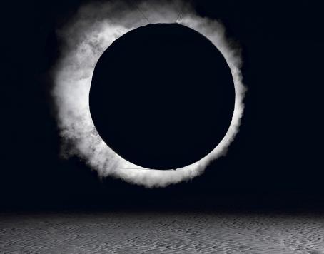 Tumman kuvan keskellä on musta ympyrä, jonka reunoilla on sumuista valoa. Ympärillä on mustaa. Maa on hiekkaista. 