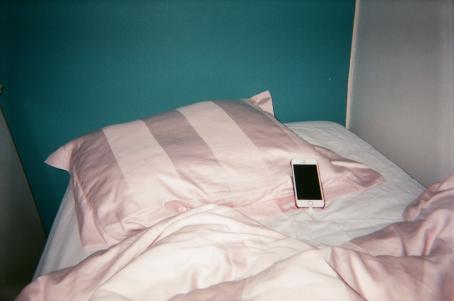 Petaamaton sänky, jossa on vaaleanpunaiset petivaatteet. Tyynyllä on puhelin. Sänky on nurkassa, ja toinen seinä on turkoosi ja toinen valkoinen. 