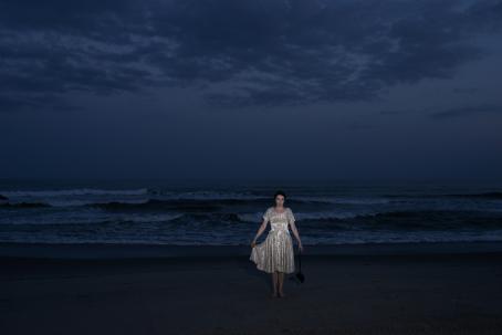 Nainen vaaleassa, kiiltävässä mekossa seisoo meren rannalla. Hän nostaa toisella kädellään mekon helmaa vähän ja toisessa kädessään hän roikottaa jotain mustaa esinettä. Naisen hahmo on valaistu salamalla, muuten kuva on tumma. 