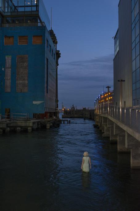 Nainen kävelee korkeiden betonirakennusten välisessä kanavassa. Vettä on lähes hänen vyötäröönsä asti. Kuva on hämärä, on luultavasti ilta.