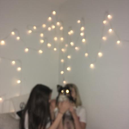 Epäterävässä kuvassa kaksi nuorta istuvat sängyllä, toinen heistä pitää koiraa sylissä. Koiran silmät näkyvät valopisteinä. Valkoisella seinällä roikkuu valosarja. 