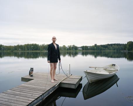 Mies seisoo laiturilla päällään pelkästään musta puvuntakki. Hän pitää toisessa kädessään köyttä, jonka toinen pää on kiinnitetty vedessä kelluvaan soutuveneeseen. Vesi on tyyni, vastarannalla näkyy vihreitä puita ja rakennuksia. Laiturilla on myös muovinen pöllö. 