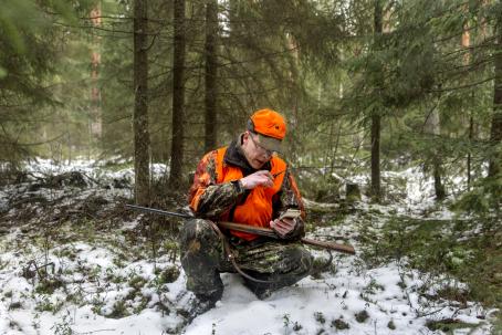 Metsästäjä kyykyssä metsässä. Hänellä on yllään maastokuvioiset housut ja takki, oranssi liivi ja lippalakki. Hän katsoo puhelinta ja puhuu radiopuhelimeen. Sylissä hänellä on kivääri. Maassa on vähän lunta.