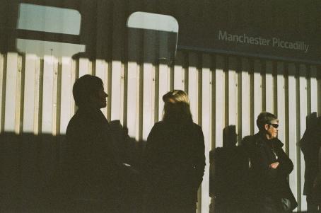 Ihmisiä seisoo seinän edessä. Osa ihmisistä jää varjoon, osa on auringossa. Ylhäällä lukee Manchester Piccadilly.