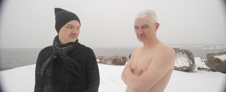 Kaksi miestä seisovat lumisella kalliorannalla, lunta sataa vaakasuoraan. Toisella miehistä on musta takki, kaulahuivi ja pipo, mutta toisella ei ole mitään vaatteita. Mies jolla on takki katsoo toista miestä, mutta paidaton mies katsoo kameraan.