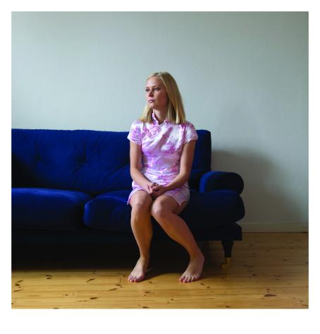 Vaaleahiuksinen nainen istuu sinisen sohvan reunalla kädet sylissään. Hänellä on yllään vaaleanpunainen kiiltävästä kankaasta tehty pitkähkö paita tai mekko, jossa on tummemmalla vaaleanpunaisella kukkien kuvia. 