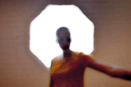 Epäterävässä kuvassa henkilö seisoo kirkkaan monikulmaisen lampun edessä. Hän ojentaa toista kättään sivulle kuvan ulkopuolelle. 
