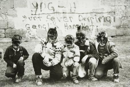 Mustavalkoisessa kuvassa viisi poikaa on osittain polvillaan, osittain kyykyssä maassa. Heidän kasvonsa on sotkettu mustalla kuvan päälle. Heidän takanaan on muuri, johon on kirjoitettu jotain. 