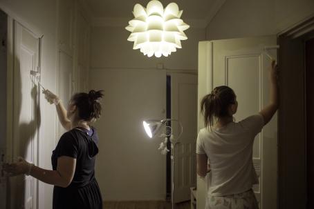 Kaksi naista maalaavat käytävän vastakkaisilla puolilla olevia ovia valkoiseksi. Molemmat naiset ovat selin kameraan. Tilaa valaisee kattolamppu ja jalkalamppu, joka osoittaa toista maalattavaa ovea.