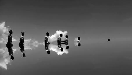 Mustavalkoisessa kuvassa ihmisiä kävelee peräkkäin kuvan poikki vasemmalta oikealle. Heidän takanaan näkyy valkoisia pilviä. Näyttää kuin kuva olisi katkaistu keskeltä ja yläosa näkyisi peilikuvana alaosassa.