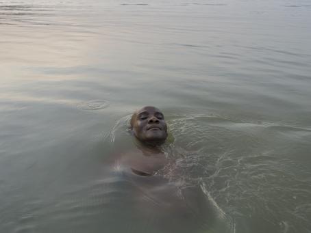 Tummaihoinen mies kelluu vedessä. Miehestä on vedenpinnan yläpuolella ainoastaan kasvot. Miehellä on silmät kiinni. 