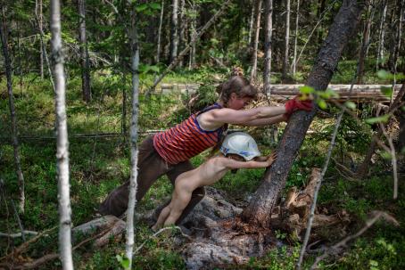 Nainen ja lapsi nojaavat puuhun kaataen sitä. Naisella on toppi ja pitkät housut, mutta lapsi on alasti, paitsi hänellä on kypärä päässään.