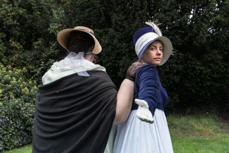 Vanhanaikaisiin mekkoihin ja hattuihin pukeutuneet kaksi naista ovat selin, toisen käsi toisen olalla. Toinen on kääntynyt kameraan päin ja ojentaa kättään. 