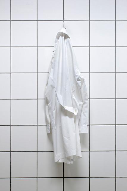Valkoisella laattaseinällä roikkuu naulassa valkoinen pitkä takki, ehkä laboratoriotakki tai lääkärintakki.