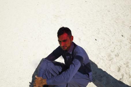 Mies istuu hiekkarannalla sinisessä kauluspaidassa ja sinisissä housuissa. Miehen päälle lankeaa osittain varjo, ja hänen kasvoilleen tulee punaista valoa. 
