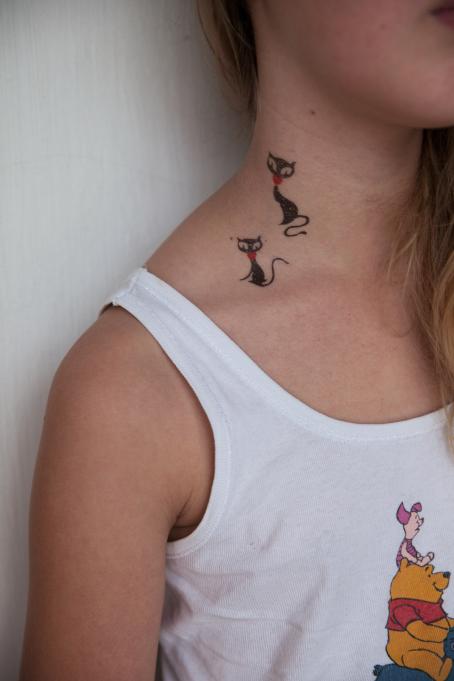 Kuvassa näkyy tytön olkapää, kaulaa ja osa kasvoista. Kaulassa on kaksi kissaa esittävää tatuointia. 