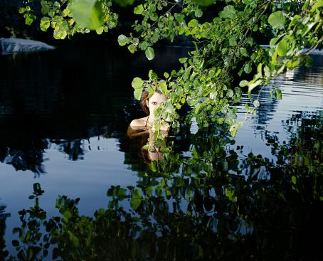 Nainen on vedessä, hänestä on pinnan yläpuolella vain hartiat ja pää. Hän jää osittain kuvan yläosasta tulevien puunoksien taakse. 