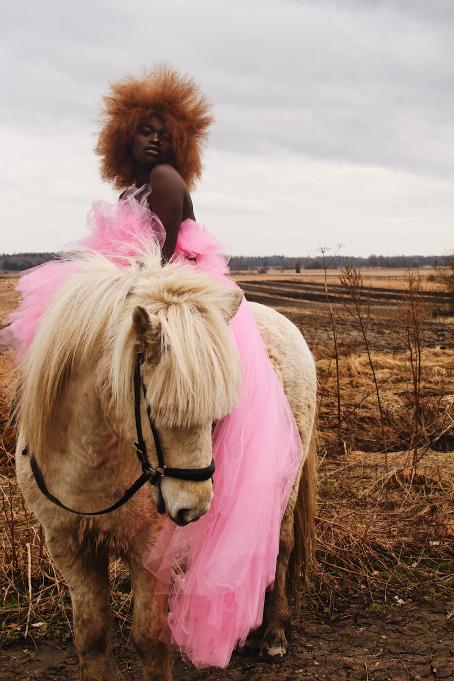 Tummaihoinen nainen istuu pienen hevosen tai ponin selässä. Hänellä on päällään vaaleanpunainen tyllihame tai -mekko, jonka helma roikkuu ponin kylkeä vasten lähes maassa asti. Naisella on vaaleanruskea afrotukka. Takana näkyy kynnettyä peltomaisemaa