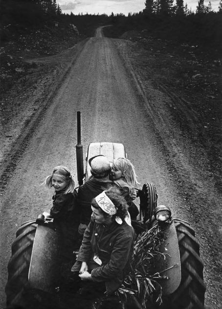 Mustavalkoisessa kuvassa neljä ihmistä istuu pienen traktorin kyydissä soratiellä. Ratissa näyttäisi olevan mies, hänen takanaan istuu nainen ja heidän ympärillään kaksi vaaleahiuksista lasta. Kuva on otettu traktorin takaa yläviistosta. 