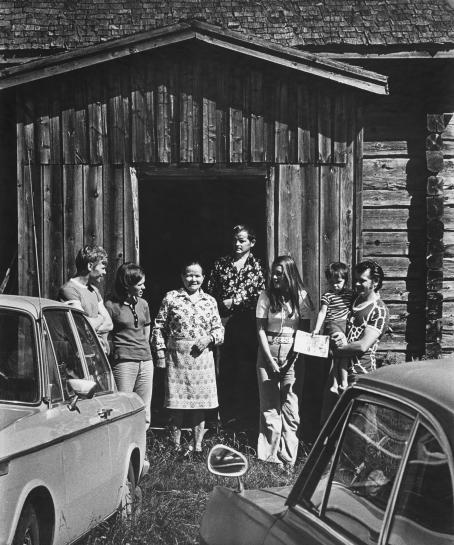Mustavalkoisessa kuvassa puutalon oven edessä seisoo seitsemän henkilöä. Keskellä on vanha nainen, hänen ympärillään nuorempia miehiä ja naisia. Yksi pitää sylissään pientä lasta. Kuvan alanurkissa näkyy kaksi autoa osittain. 